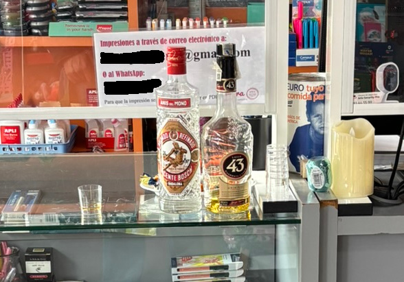 Fotografía de un mostrador en una librería del barrio. Hay una botella de Anís del Mono y otra de Licor 43 y unos vasos de chupito de plástico, para que quien quiera se pueda pegar un lingotazo de buena mañana